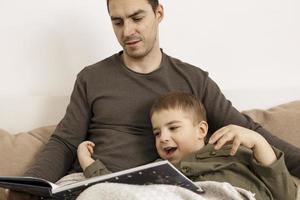 Father and son reading a book on the bed at home. Young attractive man and little boy resting in bedroom. Natural earth colors. Cozy environment. Father reads a fairy tale for his child. photo