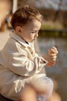 Little adorable boy sitting outdoors and eating ice cream. Lake, water and sunny weather. Child and sweets, sugar. Kid enjoy a delicious dessert. Preschool child with casual clothing. Positive emotion photo