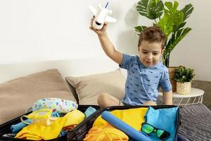 Little caucasian boy with blue shirt ready for vacation. Happy child packs clothes into a suitcase for travel. Tourist, joy of holiday. Kid at home, preparing for flying. Modern and cozy interior. photo