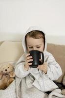 Sick little boy drinking hot tea on the bed at home. Unwell, illness child wrapped in a blanket, with mug in his room. Flu season. Interior and clothes in natural earth colors. Cozy environment. photo