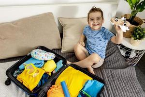 Little caucasian boy with blue shirt ready for vacation. Happy child packs clothes into a suitcase for travel. Tourist, joy of holiday. Kid at home, preparing for flying. Modern and cozy interior. photo