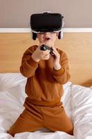 Little adorable boy sitting on bed at home with VR headset and playing interactive video game, exploring virtual reality. Cute child wearing VR glasses. Future, gadgets, technology, education online. photo