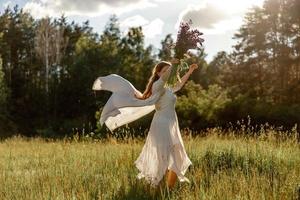 Young beautiful woman, wearing white dress, holding flowers and dancing on the meadow. Girl joying nature and freedom. Natural beauty. Dance, movement. Mental health, stress free, dreaming. Sunset. photo