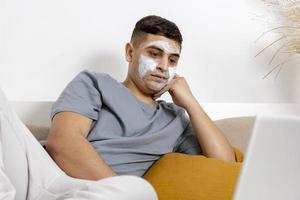 Young man with casual clothes and beauty mask on the face lying on bed with laptop computer and resting. Cosmetic for men, skin care. Man reading news in internet or watching movie. Relaxing at home. photo