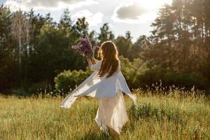 Young beautiful woman, wearing white dress, holding flowers and dancing on the meadow. Girl joying nature and freedom. Natural beauty. Dance, movement. Mental health, stress free, dreaming. Sunset. photo