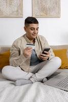 joven sonriendo mientras navega por la web en su teléfono inteligente y tiene una tarjeta de crédito en la mano tratando de comprar algo en línea. compras en línea fáciles en casa, comercio electrónico. interior acogedor. foto