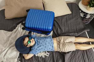 Little caucasian boy with blue shirt ready for vacation. Happy child with blue suitcase going to travel. Tourist, joy of holiday. Kid at home, preparing for flying. Modern and cozy interior. photo