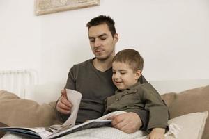 Father and son reading a book on the bed at home. Young attractive man and little boy resting in bedroom. Natural earth colors. Cozy environment. Father reads a fairy tale for his child. photo