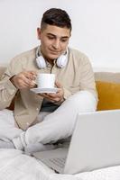 Young man with casual clothes sitting on bed with laptop computer and drinking tea or coffee. Man using notebook to surf in internet, read news, watch movie, call friends. Relaxing at home. photo