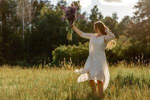 Young beautiful woman, wearing white dress, holding flowers and dancing on the meadow. Girl joying nature and freedom. Natural beauty. Dance, movement. Mental health, stress free, dreaming. Sunset. photo