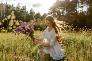 joven mujer hermosa, vestida de blanco, sosteniendo flores en el prado. chica disfrutando de la naturaleza y la libertad. belleza natural. libre de estrés, soñando. puesta de sol. foto