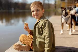 Little adorable boy sitting outdoors and eating ice cream. Lake, water and sunny weather. Child and sweets, sugar. Kid enjoy a delicious dessert. Preschool child with casual clothing. Positive emotion photo