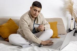 Young man with casual clothes surfing the web on his laptop computer and holding credit card in his hand trying to buy something online. Easy online shopping at home, e-commerce. Cosy interior. photo