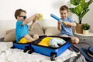 Two little boys with blue shirts ready for vacation. Happy children packing clothes into a suitcase for travel. Tourist, joy of holiday. Kids at home, preparing for flying. Modern and cozy interior. photo