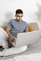 Young man with casual clothes and beauty mask on the face lying on bed with laptop computer and resting. Cosmetic for men, skin care. Man reading news in internet or watching movie. Relaxing at home. photo