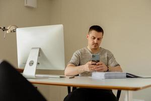 Young caucasian man sitting at his desk in the office and working with computer and smartphone, answering emails. Young employee has a working day in office. photo