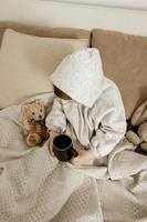 Sick little boy drinking hot tea on the bed at home. Unwell, illness child wrapped in a blanket, with mug in his room. Flu season. Interior and clothes in natural earth colors. Cozy environment. photo