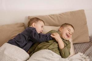 Two little and cute caucasian boys playing together on the bed at home. Interior and clothes in natural earth colors. Cozy environment. Children having fun, two brothers kittle each other. photo