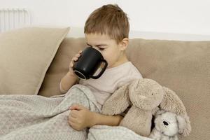Sick little boy drinking hot tea on the bed at home. Unwell, illness child wrapped in a blanket, with mug in his room. Flu season. Interior and clothes in natural earth colors. Cozy environment. photo