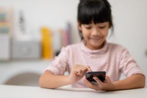 Cute little girl uses smartphone while sitting at the sofa in the living room. Child surfing the internet on mobile phone, browses through internet and watches cartoons online at home photo