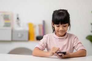 Cute little girl uses smartphone while sitting at the sofa in the living room. Child surfing the internet on mobile phone, browses through internet and watches cartoons online at home photo