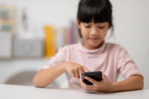 Cute little girl uses smartphone while sitting at the sofa in the living room. Child surfing the internet on mobile phone, browses through internet and watches cartoons online at home photo