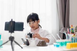 Asian school girl making easy chemistry experiments and recording a video for his followers, Young blogger kid posing in front of camera for vlog, Children make vlog for social media channel concept. photo