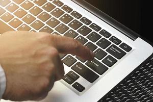 Businesssman hand touching enter button on Laptop keyboard with sun ray effect. Computer notebook with man index finger push keypad. Closeup of laptop computer with forefinger for business concept. photo