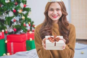 retrato de una mujer joven con cara hermosa y sonrisa dando regalos de navidad dentro de la casa con un árbol de navidad verde decorado con bombillas y cajas de regalo para el intercambio. concepto del día de navidad foto