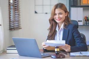 Attractive smiling attractive lawyer in a black suit reading case documents to a client in the office with a laptop computer with a lawyer hammer on the desk. Ideas for attorneys accepting lawsuits photo