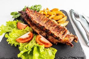 a large kind of pork ribs on a slate board with lettuce greens, slices of tomato and grilled potatoes. the concept of food cooked in the backyard. photo