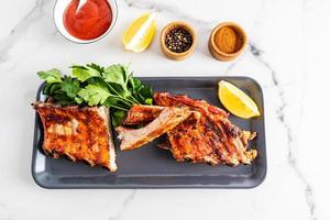 Grilled pork ribs are served on a rectangular dark ceramic plate with fresh vegetables and a bowl of sauce. white background. recipe menu. photo