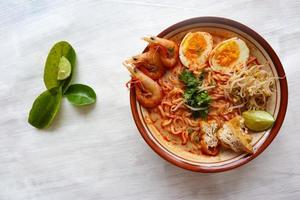 laksa, curry laksa elaborado con fideos, fideos, brotes de soja, huevo, tofu, lima y camarones con una salsa de sopa picante rica en especias. laksa servido en tazón, aislado sobre fondo blanco. foto