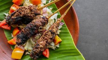 sate buntel o satay buntel es satay hecho de cordero picado envuelto en grasa de cordero y luego asado a la parrilla. condimento con tomates, chalotes, pimienta, repollo foto