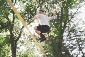 Young man balancing and jumping on slackline. Man walking, jumping and balancing on rope in park. photo