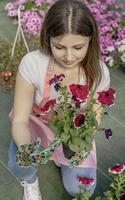 Young woman at a nursery holding flower plant in her hands as she kneels in the walkway between plants. photo