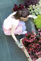 Young woman at a nursery holding flower plant in her hands as she kneels in the walkway between plants. photo