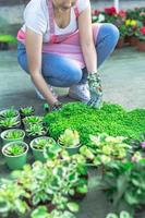 Young woman at a nursery holding flower plant in her hands as she kneels in the walkway between plants. photo