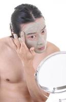 Beauty procedures skin care concept. Young man applying facial gray mud clay mask on his face photo
