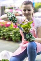 Young woman at a nursery holding flower plant in her hands as she kneels in the walkway between plants. photo