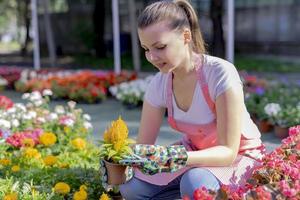 Young woman at a nursery holding flower plant in her hands as she kneels in the walkway between plants. photo