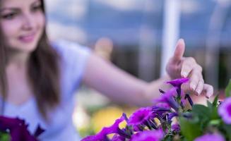 mujer joven en un vivero sosteniendo una planta de flores en sus manos mientras se arrodilla en el camino entre las plantas. foto