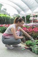 Young woman at a nursery holding flower plant in her hands as she kneels in the walkway between plants. photo