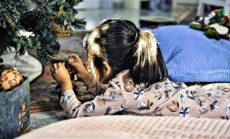 Asian little girl decorate the Christmas tree indoors. The morning before Xmas. Portrait loving girl close up. photo