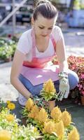 Young woman at a nursery holding flower plant in her hands as she kneels in the walkway between plants. photo