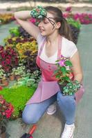 mujer joven en un vivero sosteniendo una planta de flores en sus manos mientras se arrodilla en el camino entre las plantas. foto