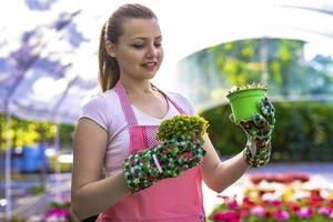 mujer joven en un vivero sosteniendo una planta de flores en sus manos mientras se arrodilla en el camino entre las plantas. foto