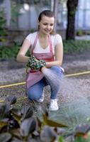 mujer joven en un vivero sosteniendo una planta de flores en sus manos mientras se arrodilla en el camino entre las plantas. foto