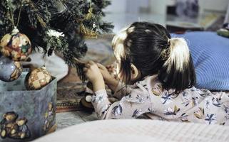 Asian little girl decorate the Christmas tree indoors. The morning before Xmas. Portrait loving girl close up. photo