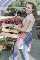 Young woman at a nursery holding flower plant in her hands as she kneels in the walkway between plants. photo